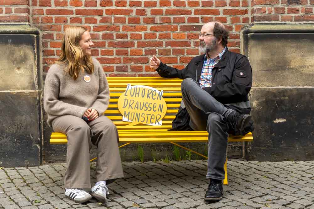Sara Stetter und Dr. Achim Detering sitzen ab Juni als ehrenamtliche Zuhörerin und ehrenamtlicher Zuhörer auf den gelben Bänken in Münster