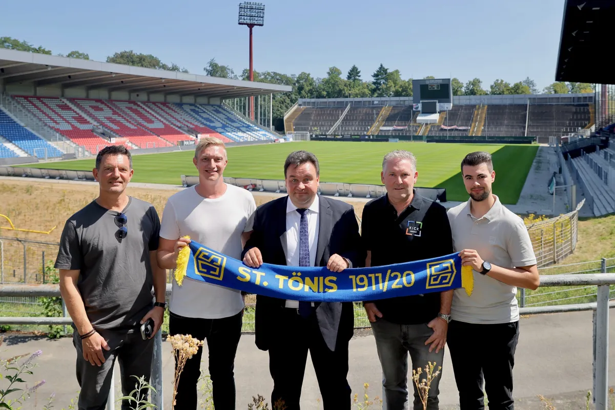 Fünf Männer stehen bei sonnigem Wetter vor der Tribüne und dem Spielfeld der Krefelder Grotenburg. Drei von ihnen halten gemeinsam einen blau-gelben Fanschal mit der Aufschrift „St. Tönis 1911/20“ in die Kamera